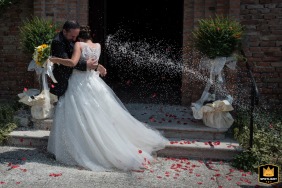 In Malcantone, Mantova, Italy, the newlyweds embrace joyfully as they exit the ceremony. Surrounded by friends and family, they are showered with a final burst of rice.